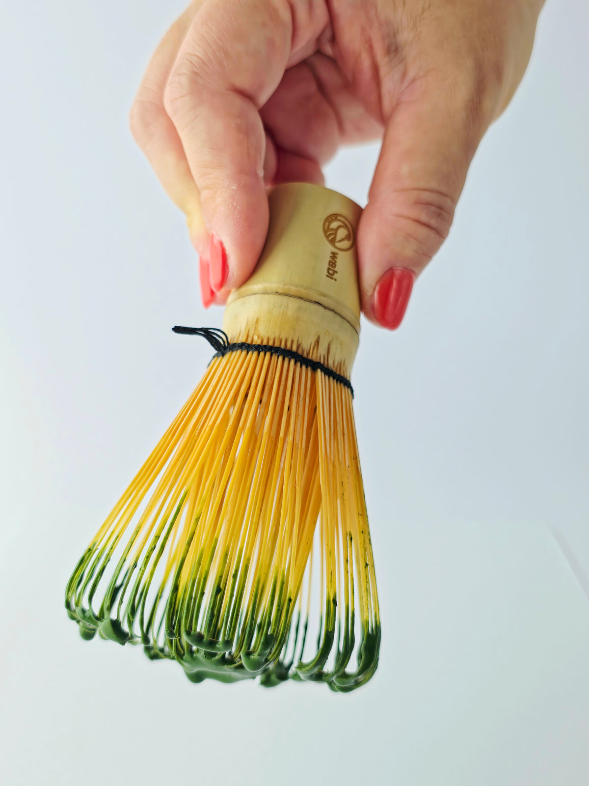 Wabi Matcha Eiko Ceremonial matcha on a bamboo whisk held in a woman's hand with red nails and a white background.