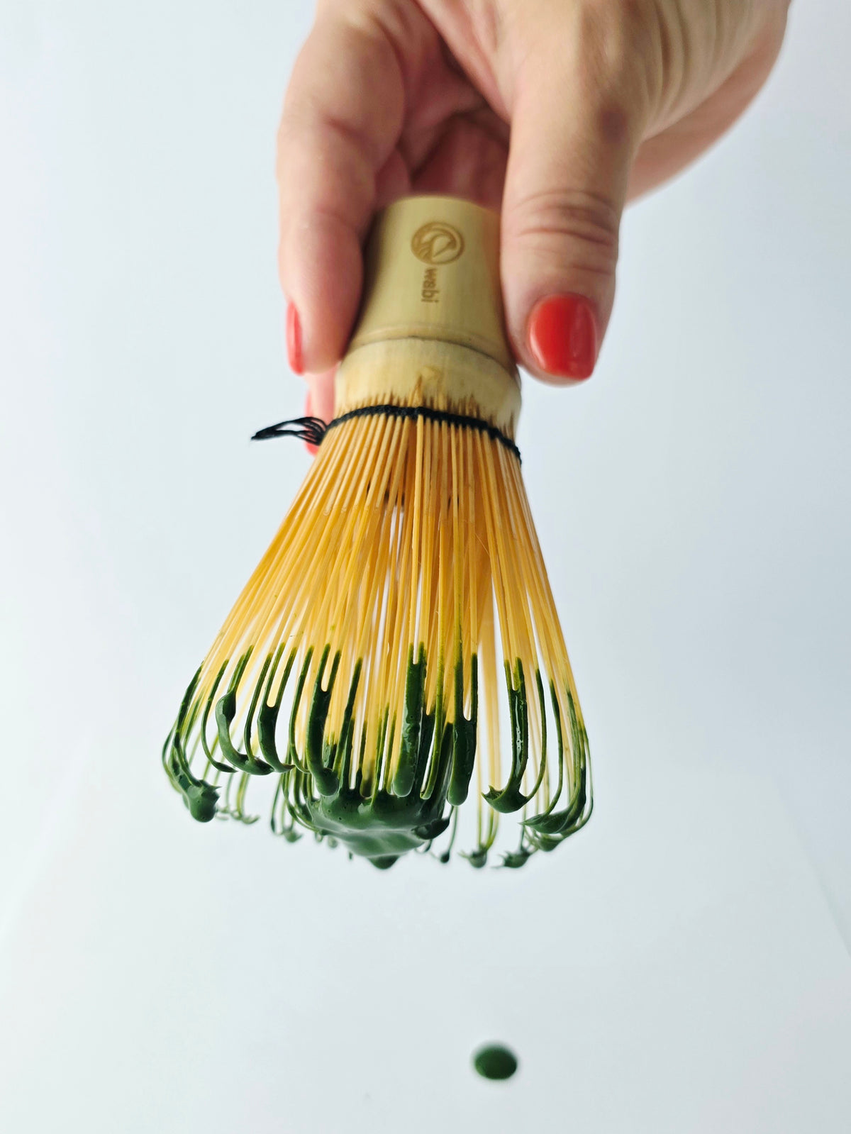 Hand holding a traditional Japanese tea whisk against a white background with matcha dripping from the prongs