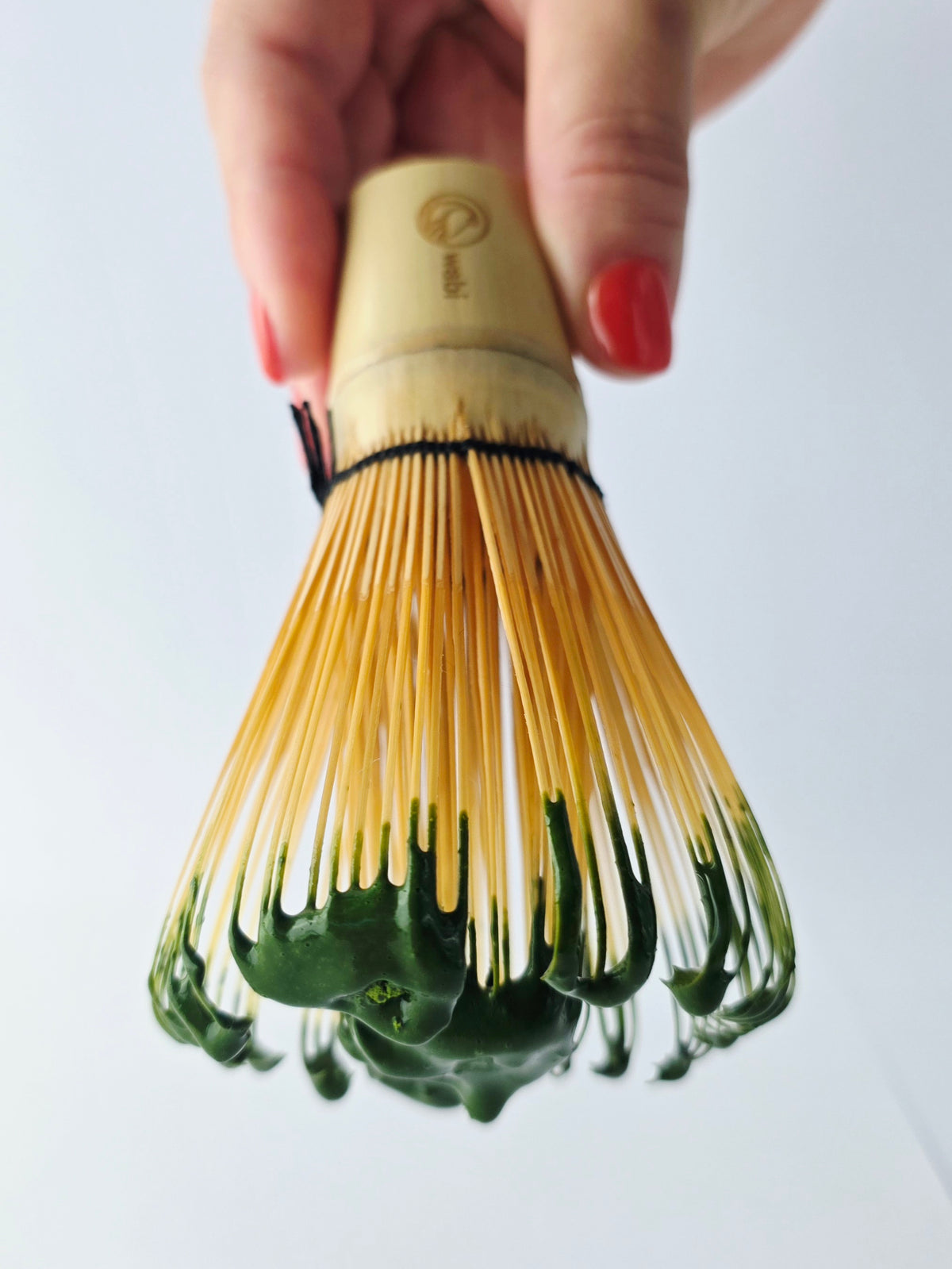 Bamboo whisk with green tea leaves held by a hand on a white background with matcha dripping from prongs.