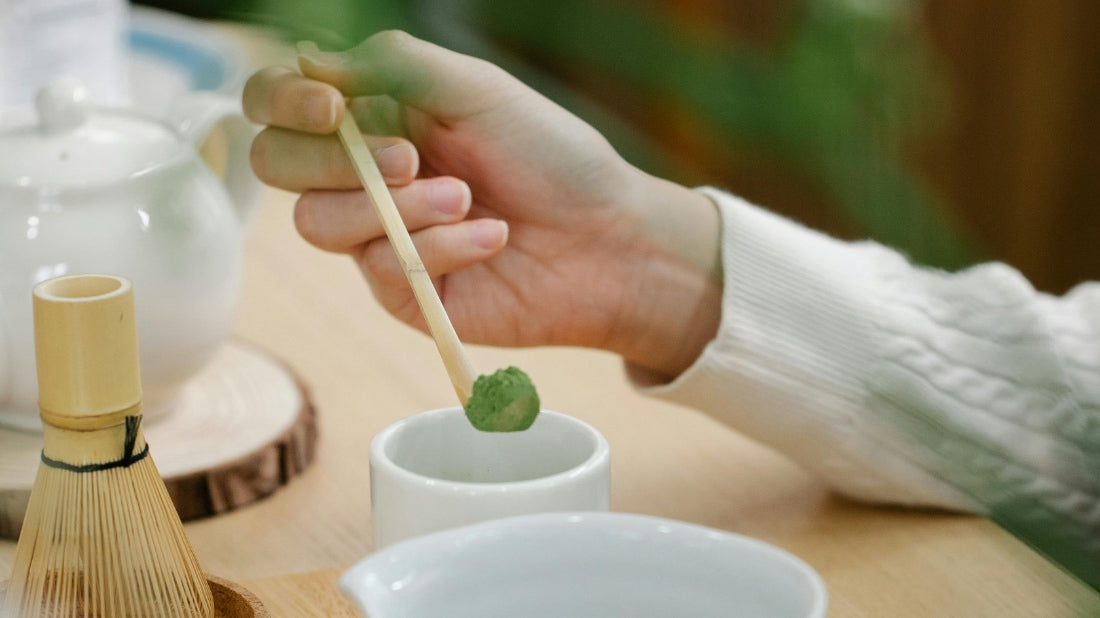 An ladies hand placing a serving of matcha powder on a bamboo spoon being placed into a white matcha bowl.