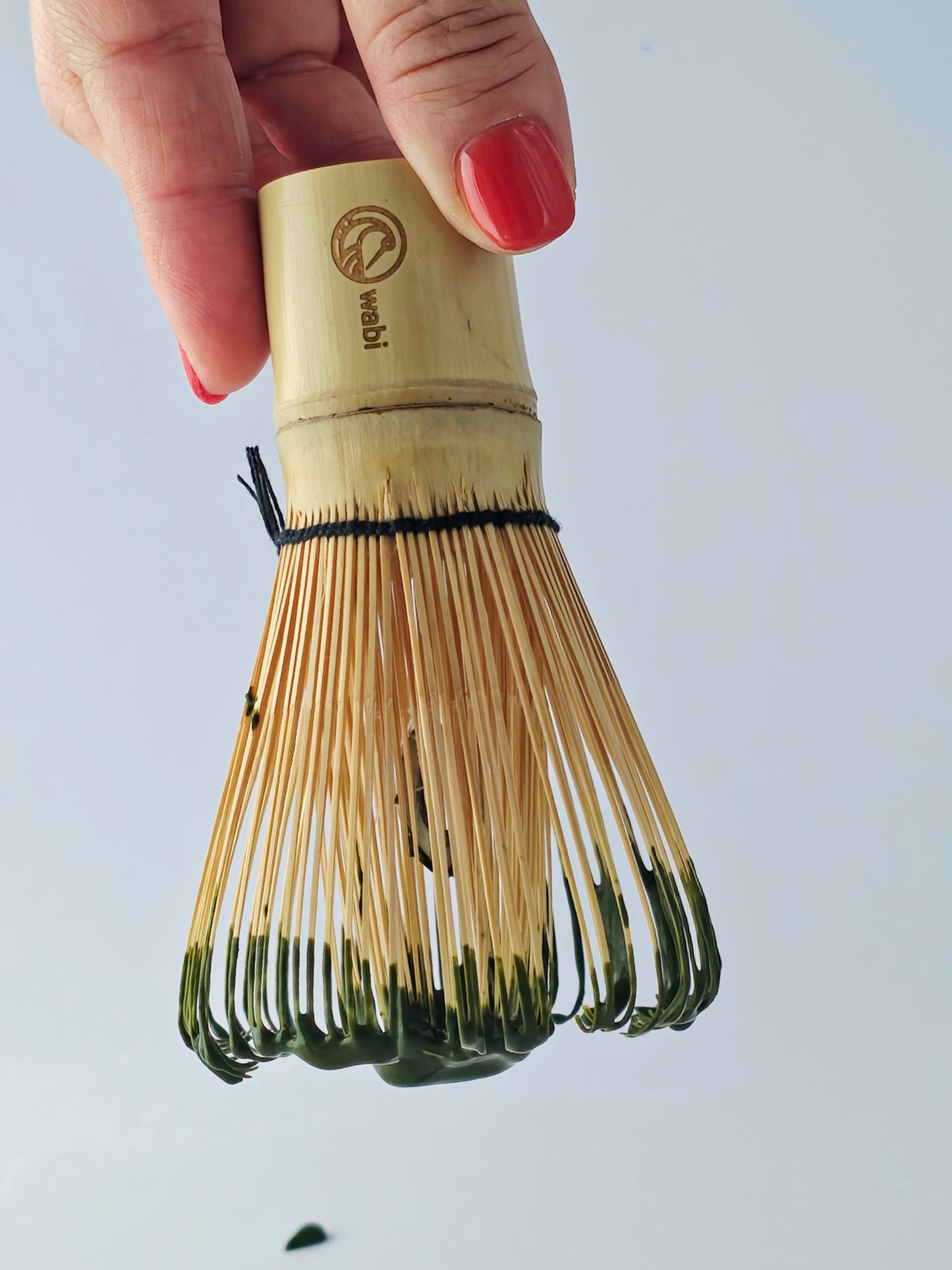 Bamboo whisk held by a hand with red nail polish against a light gray background