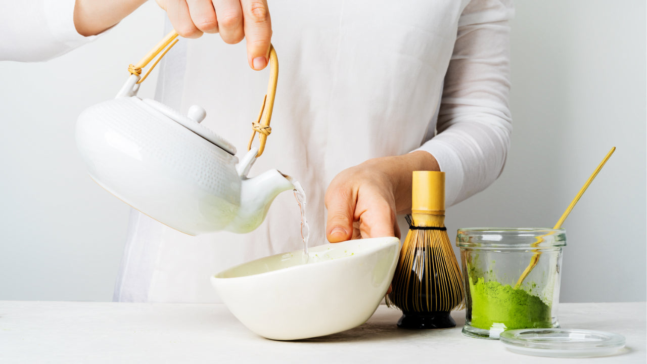 A person wearing white pouring hot water from a white jug with a bamboo whisk and jar of bright green matcha with a bamboo scoop sitting in it.